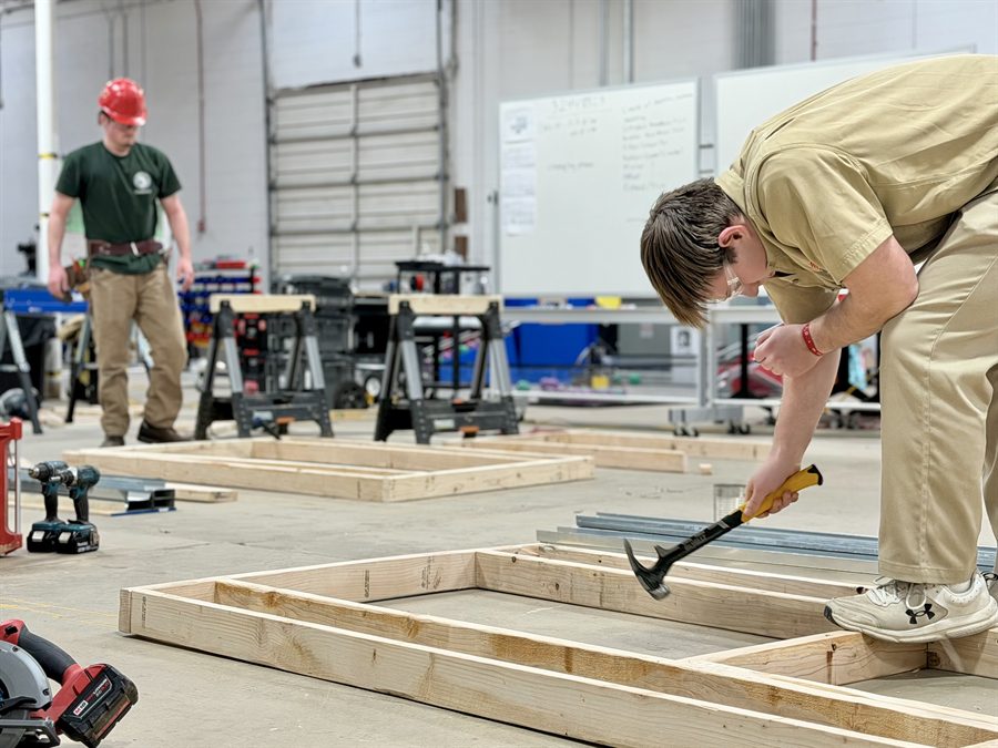 Photo of a man using a hammer on a wooden frame with another man in the back wearing a construction helmet.