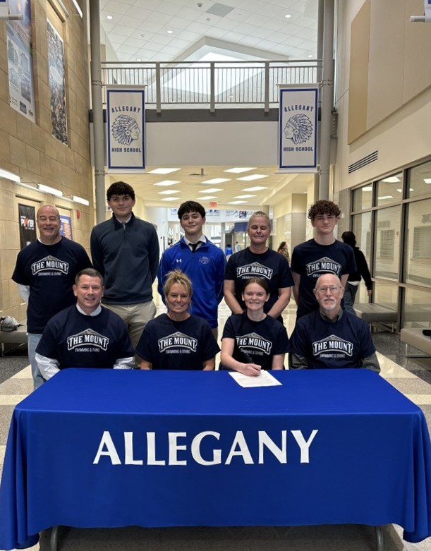 Photo of a student signing to a college for athletics with multiple people sitting around here and some standing behind her.