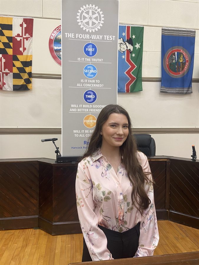 Photo of student standing in front of a banner about 'The Four Way Test." On the wall behind hangs multiple flags.