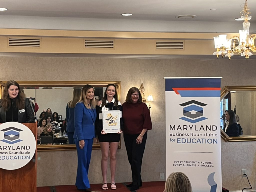 Photo of a student standing in the center of two women, holding a certificate. To the left is a woman at a podium and to the right is a standing banner with info on Maryland Business Roundtable for Education.