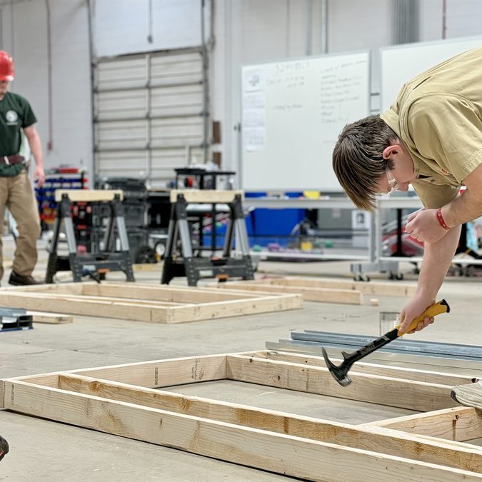 Photo of a man using a hammer on a wooden frame with another man in the back wearing a construction helmet.