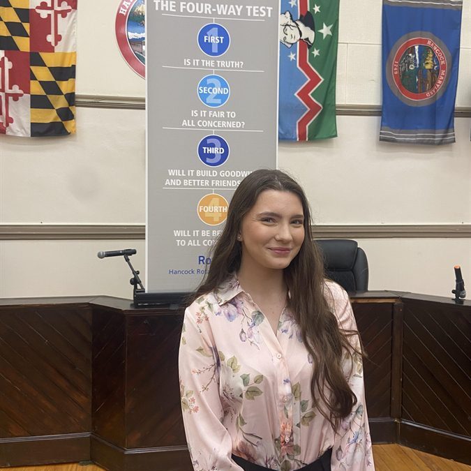 Photo of student standing in front of a banner about 'The Four Way Test." On the wall behind hangs multiple flags.